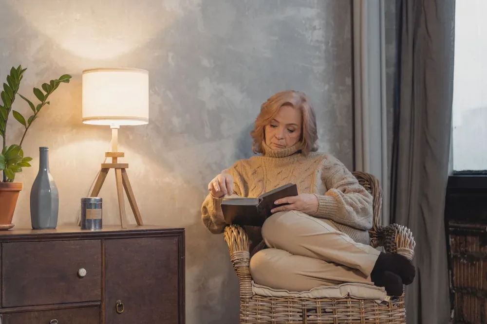 Femme mature assise dans un fauteuil en osier, lisant un livre chez elle