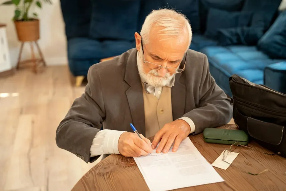 Un homme âgé signe un document assis à une table.