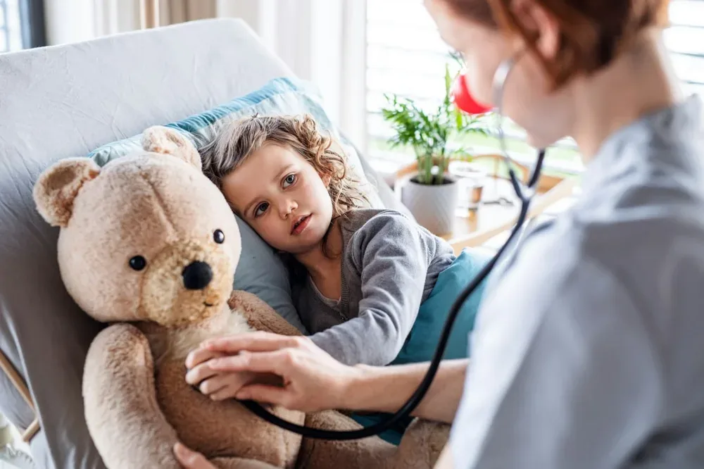 Une médecin pose son stéthoscope sur un ours en peluche auprès d'une petite fille allongée.