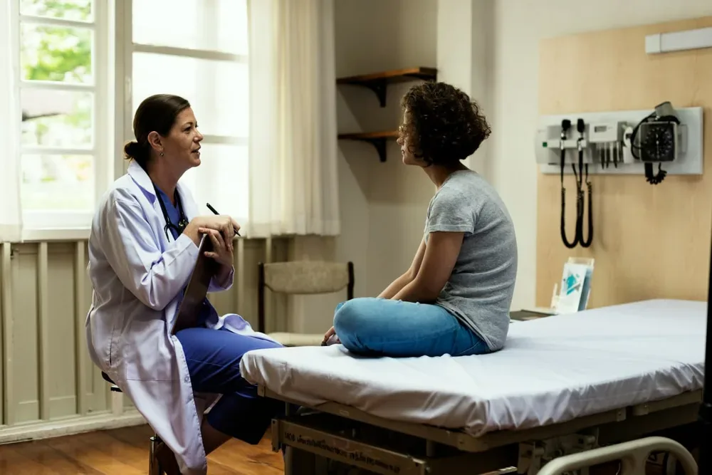 Une professionnelle de santé échange avec une femme assise sur une table d’examen dans un cabinet médical lumineux.